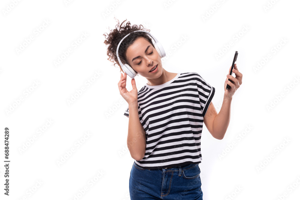 young well-groomed curly woman with black hair in a summer t-shirt listens to music in wireless headphones and holds a phone young well-groomed curly woman with black hair in a summer t-shirt listens to music in wireless headphones and holds a phone