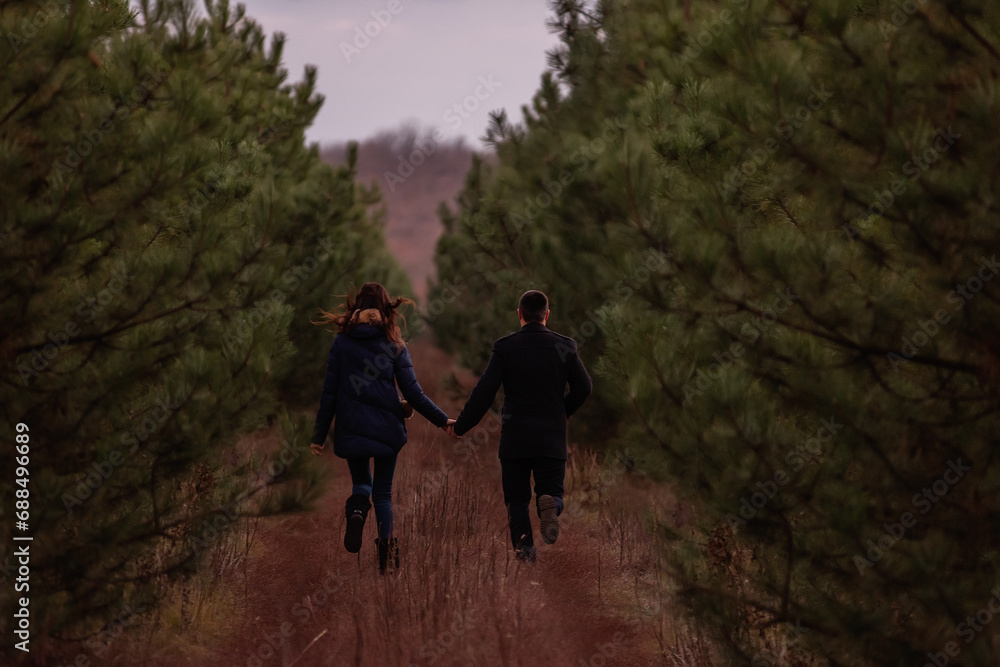 Obraz premium Happy couple running through forest holding hands. Man in coat and a young woman are fooling around