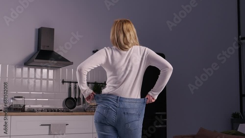 Rear view of slim elegant mature woman wearing too large old pants, showing results and achievement after successful slimming program while standing in domestic room. photo