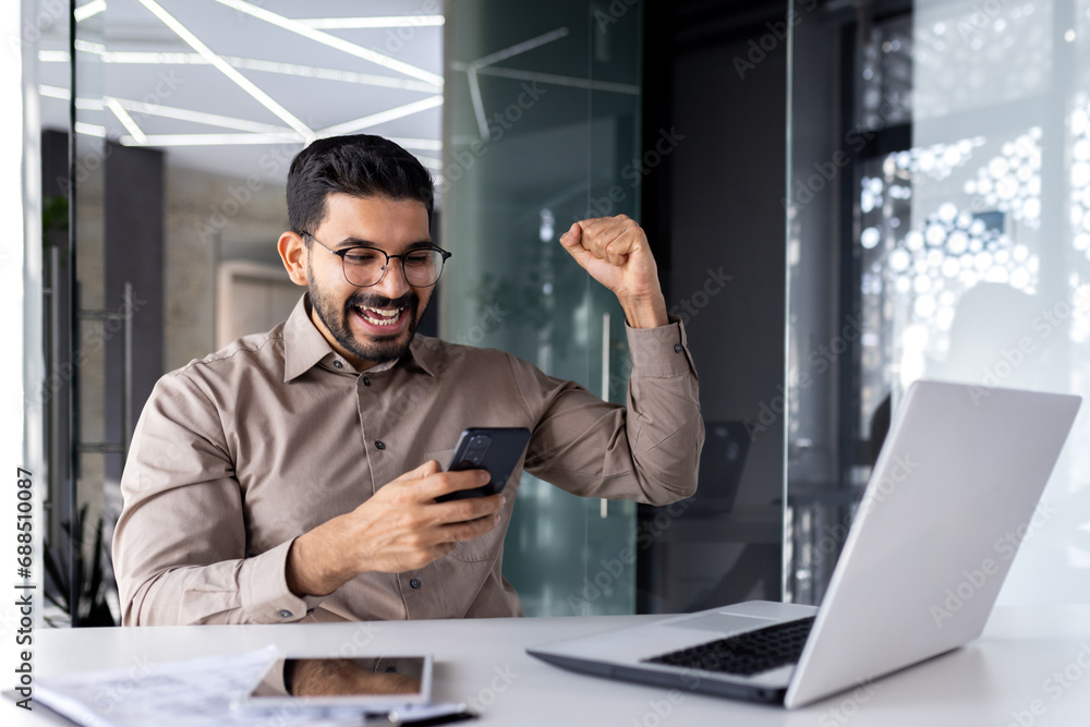 Successful businessman winner inside office at workplace, man received online notification of victory win, worker at desk with laptop celebrating triumph