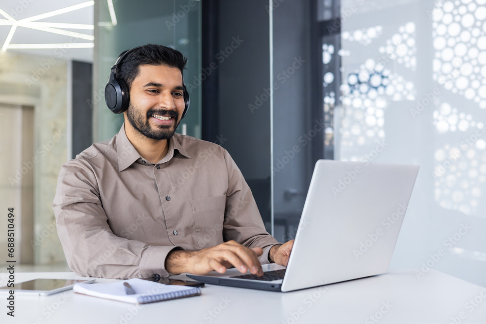 Young successful Indian programmer at workplace inside office, man with headphones smiling satisfied, coding new software using laptop at table, businessman in business shirt successful and satisfied Young successful Indian programmer at workplace inside office, man with headphones smiling satisfied, coding new software using laptop at table, businessman in business shirt successful and satisfied