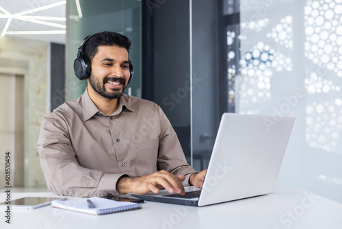 Young successful Indian programmer at workplace inside office, man with headphones smiling satisfied, coding new software using laptop at table, businessman in business shirt successful and satisfied