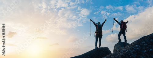 Silhouette of Asian Male and female standing raised hands with trekking poles on cliff edge on top of rock mountain with beautiful sunset background, Asia couple hiking in sunset background.
