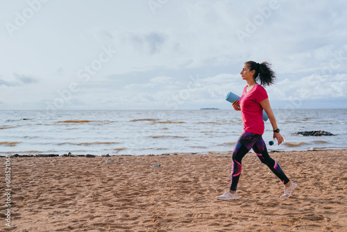 A fit Latin American girl in sportswear sneakers carries a sports mat in her hands walks along the beach going to work out meditate and practice yoga against view in ocean. Outdoor exercise.