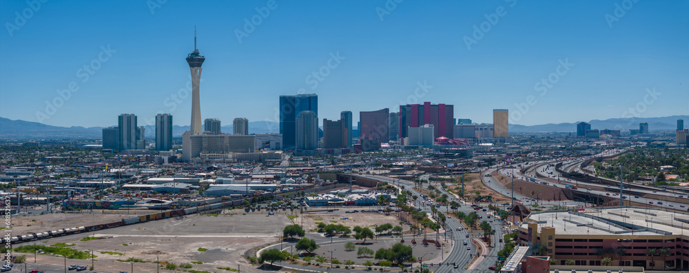 Aerial view of Las Vegas, Nevada, highlighting the Stratosphere Tower amid a bustling cityscape with green spaces, under a clear blue sky with mountains beyond.