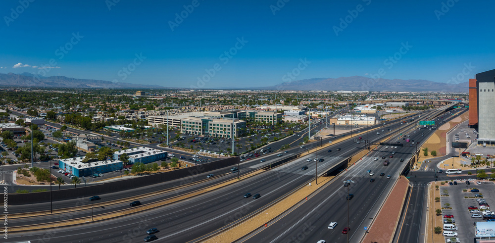 Aerial view of a bustling Las Vegas cityscape with a busy highway, surrounded by modern beige buildings, green spaces, and mountains under a clear blue sky.