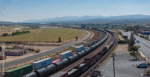 Elevated view of a long freight train traversing Barstow's scenic landscape, with clear skies, sparse vegetation, distant mountains, and a bustling road foreground.