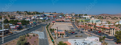 Aerial view of a Barstow U.S. town nestled in a desert valley, featuring beige and brown buildings with terracotta roofs, palm trees, and a grid-like street layout.