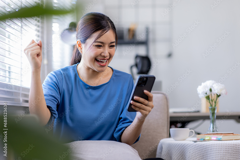 Young Happy Asian woman smiling holding Phone with fist hand and excited for success on sofa at home.