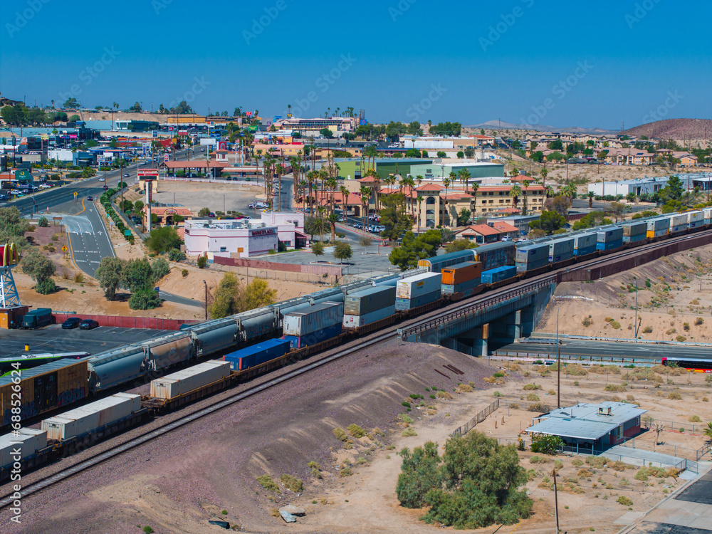 Panoramic view of a colorful freight train traversing a semi-arid Southwestern landscape with a Barstow town backdrop, clear skies, and Route 66 charm.
