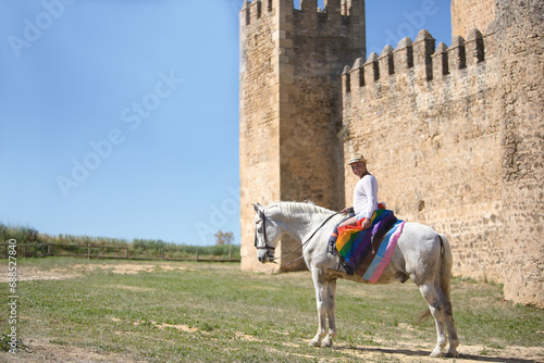Non-binary person, young and South American, very makeup, mounted on a white horse, smiling and happy, with a gay pride flag on the rump, next to an old medieval castle. Concept queen, lgbtq+, pride.