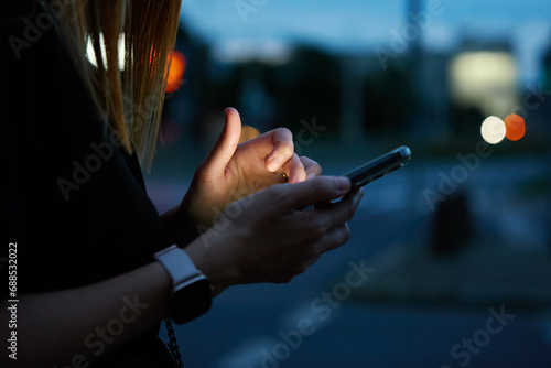 Close up shot of woman using smartphone on city street with bokeh lights at night. Mobile phone in female hands outdoors photo