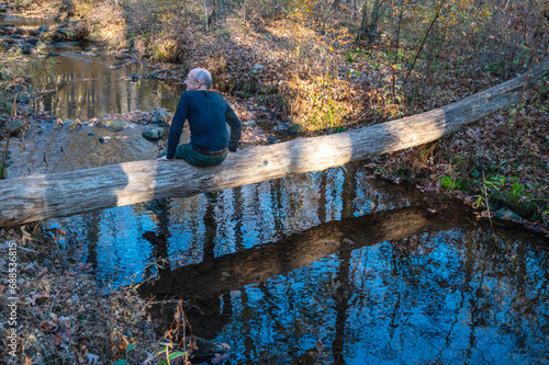 Senior man sits alone on fallen log footbridge over stream profile