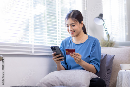 Mobile Shopping. Cheerful Asian Woman Using Smartphone Shopping Online Holding Credit Card Making Payment Sitting At Sofa At Home. Internet Banking Application And E-Commerce

