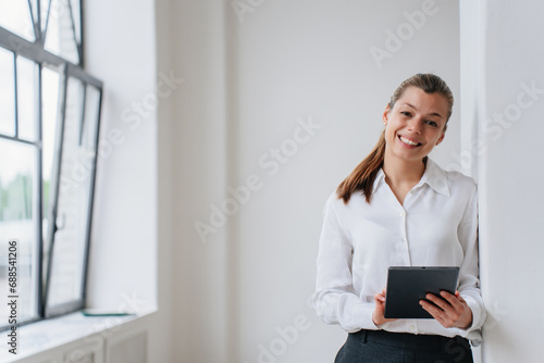 Cheerful student girl thinks about future, standing indoor, leans on wall looks aside holds tablet. Attractive thoughtful hispanic woman designer stands against empty interior. Successful people