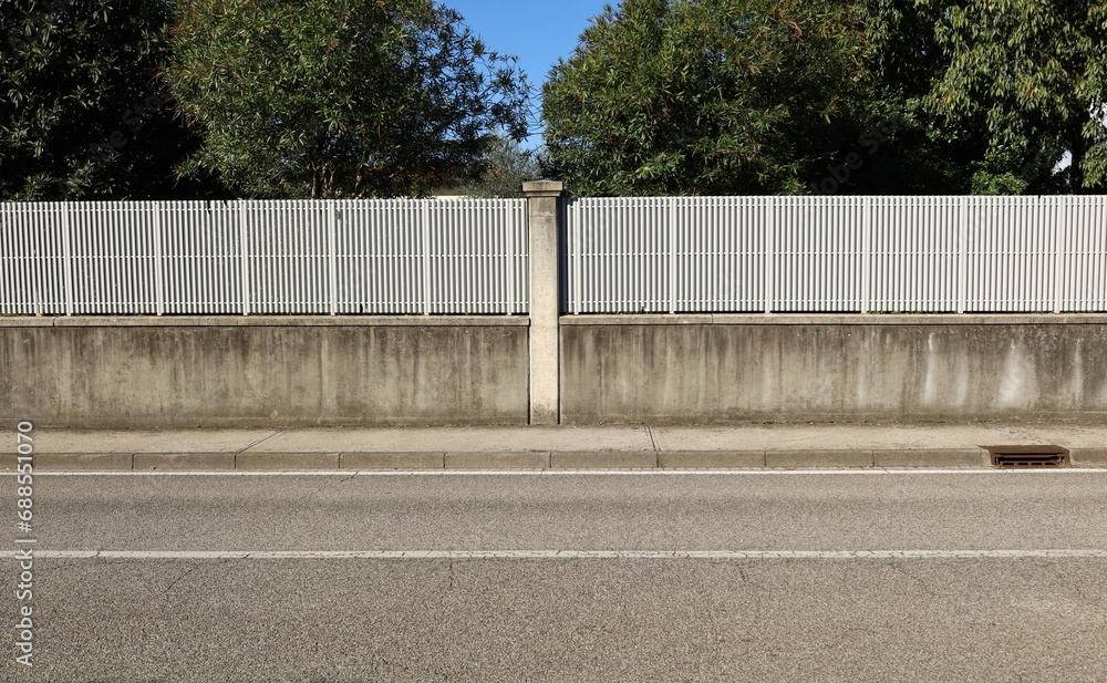 Fence made of concrete wall with white railing on top. Decorative column in the middle. Trees on behind. Cement tiled sidewalk and street in front. Background for copy space. Fence made of concrete wall with white railing on top. Decorative column in the middle. Trees on behind. Cement tiled sidewalk and street in front. Background for copy space.