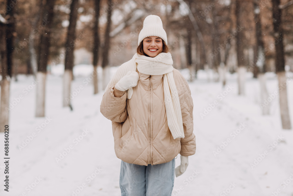 Happy smiling young woman dressed down jacket scarf hat and mittens enjoys winter weather and walking through the snowy winter park Happy smiling young woman dressed down jacket scarf hat and mittens enjoys winter weather and walking through the snowy winter park
