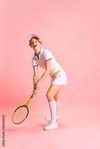 Happy cheerful young lady, dressed retro tennis uniform with racquet getting high from workout against pastel pink background © Lustre