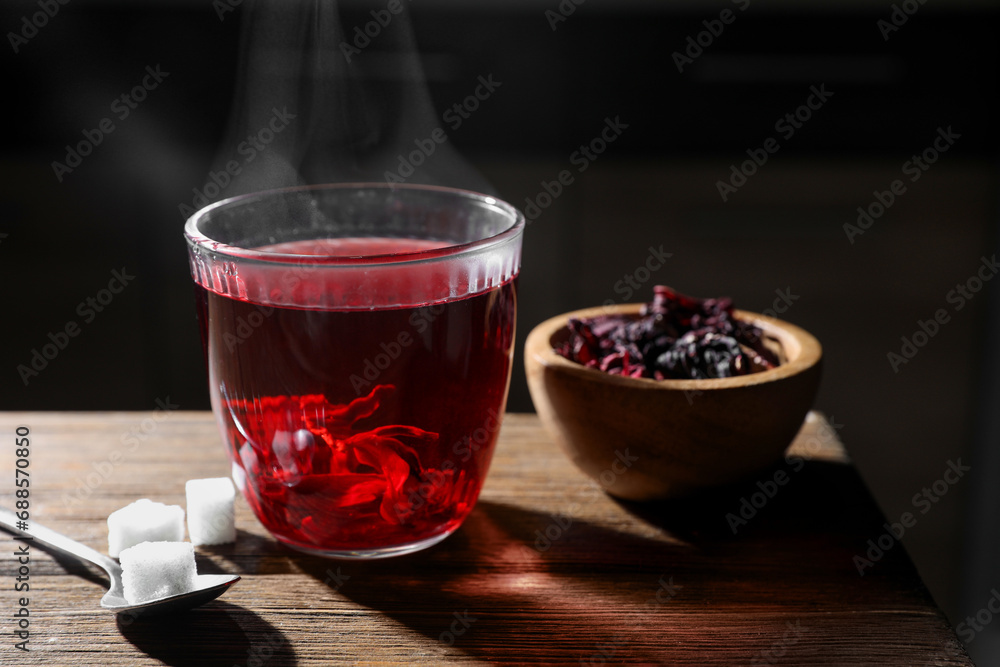 Delicious hibiscus tea in glass, sugar cubes and dry roselle petals on wooden table, closeup Delicious hibiscus tea in glass, sugar cubes and dry roselle petals on wooden table, closeup