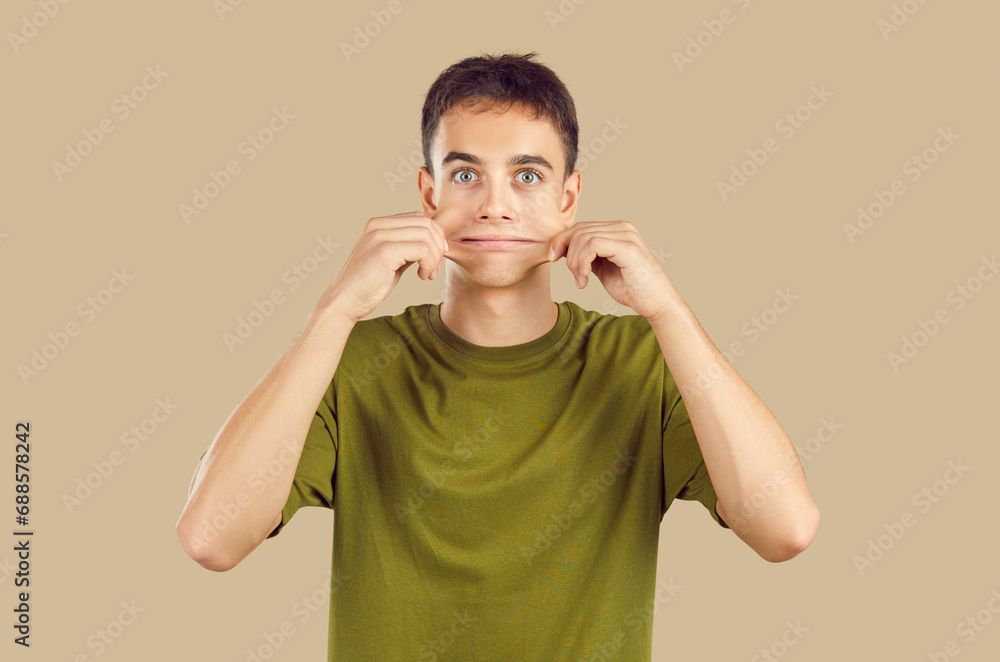 Close up studio portrait of young grimacing boy student wearing casual green t-shirt having fun pulling back his cheeks isolated on a beige background. People emotions, facial expression concept. - obrazy, fototapety, plakaty 