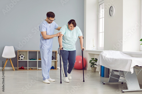 Male doctor helps female patient with leg injury. Man in scrubs uniform supports fat overweight woman while she walks with crutches during bone and muscle rehabilitation in physiotherapy center