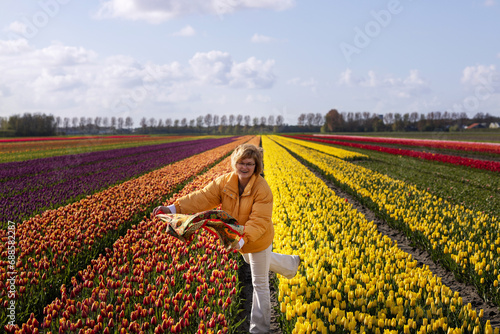 A young woman in a yellow jacket catches a kerchief torn off by the wind in a field with colorful tulips
