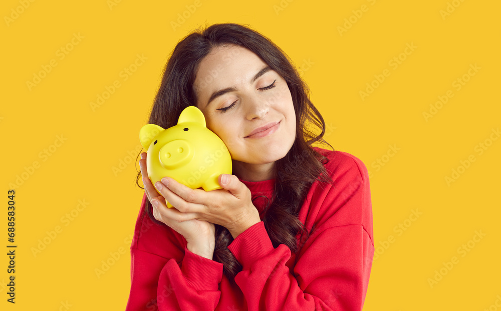 Smiling young woman gently leans her cheek to piggy bank, isolated on orange background. Saving, finances, banking, investment concept for banner, website design or landing web page. Smiling young woman gently leans her cheek to piggy bank, isolated on orange background. Saving, finances, banking, investment concept for banner, website design or landing web page.
