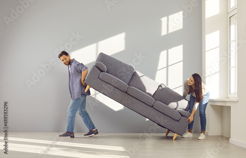 Happy young couple carrying sofa to their new home. Cheerful husband and wife placing sofa in empty sunny living room. Family moving in new apartment or remodeling home photo