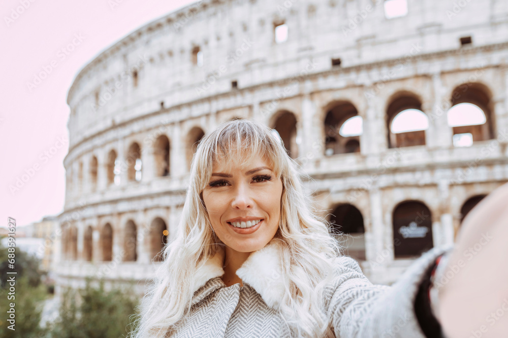 Smiling young beautiful woman tourist taking selfie during autumn, winter, at Rome Colosseum, Italy. Vacations, travel, trip concept.