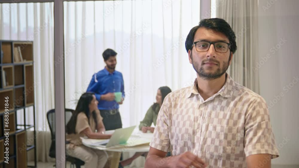 A Modern young Indian male corporate employee wearing eyeglasses with arms crossed looking at the camera confidently with busy office coworkers interacting with each other in the background. A Modern young Indian male corporate employee wearing eyeglasses with arms crossed looking at the camera confidently with busy office coworkers interacting with each other in the background.