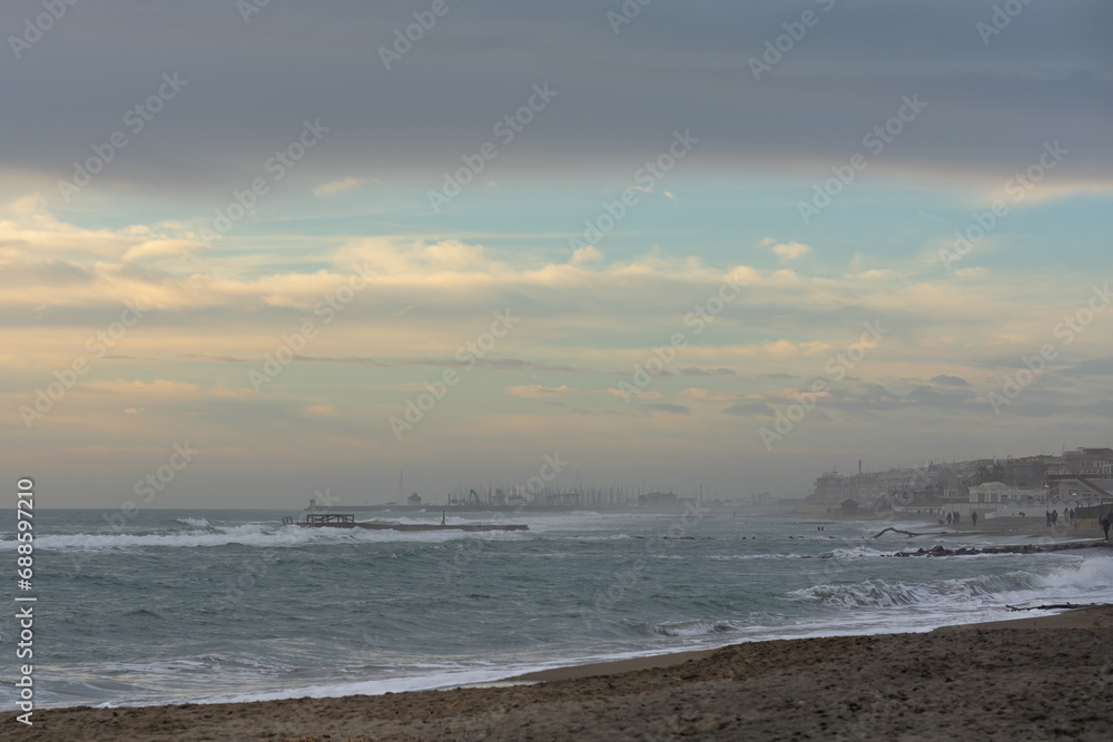 Strong gale force winds on the Mediterranean coast, a view of the coastline, buildings and people walking in the distance. Gray-white clouds in the sky with blue gaps Strong gale force winds on the Mediterranean coast, a view of the coastline, buildings and people walking in the distance. Gray-white clouds in the sky with blue gaps