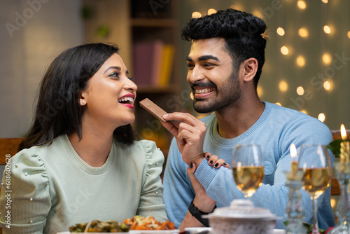 Happy boyfriend giving chocolate byte to girl friend during candle light dinner at restaurant - concept of valentine's day celebration, romantic couples and intimacy.