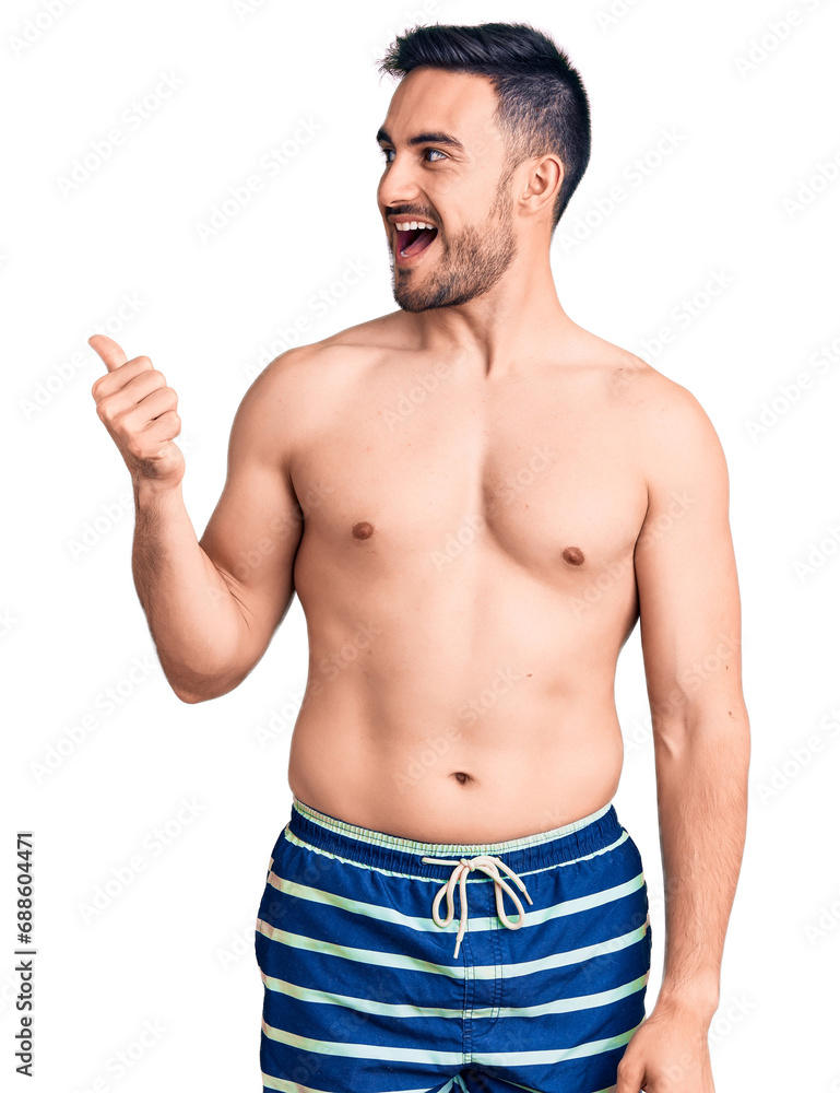 Young handsome man wearing swimwear smiling with happy face looking and pointing to the side with thumb up. Young handsome man wearing swimwear smiling with happy face looking and pointing to the side with thumb up.