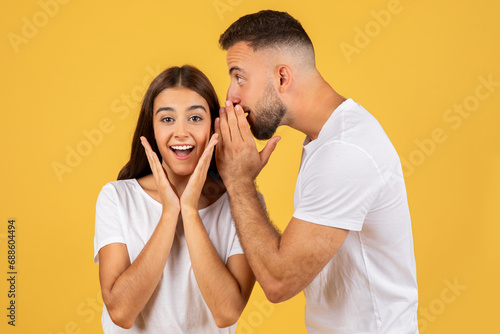 Happy young european man in white t-shirt whispers in ear of surprised lady secret