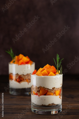 Parfait in a glasses with persimmon, rosemary, pecan, whipped cream and biscuit on a brown. Healthy food, vegan, sugar, gluten and lactose free.