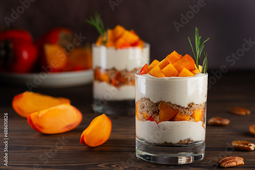 Dessert in a glasses with persimmon, rosemary, pecan, whipped cream and biscuit on a wooden table. Healthy food, vegan, sugar, gluten and lactose free.