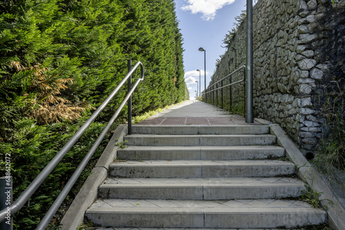Fototapeta Naklejka Na Ścianę i Meble -  Stairs with metal railing on a narrow street flanked by a wall and a large unpruned hedge