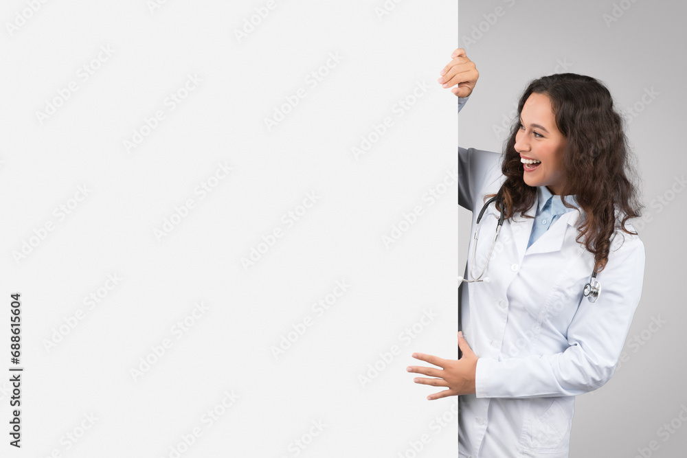 Cheerful young female doctor peeking from behind blank white placard board for text or design, free space, banner Cheerful young female doctor peeking from behind blank white placard board for text or design, free space, banner