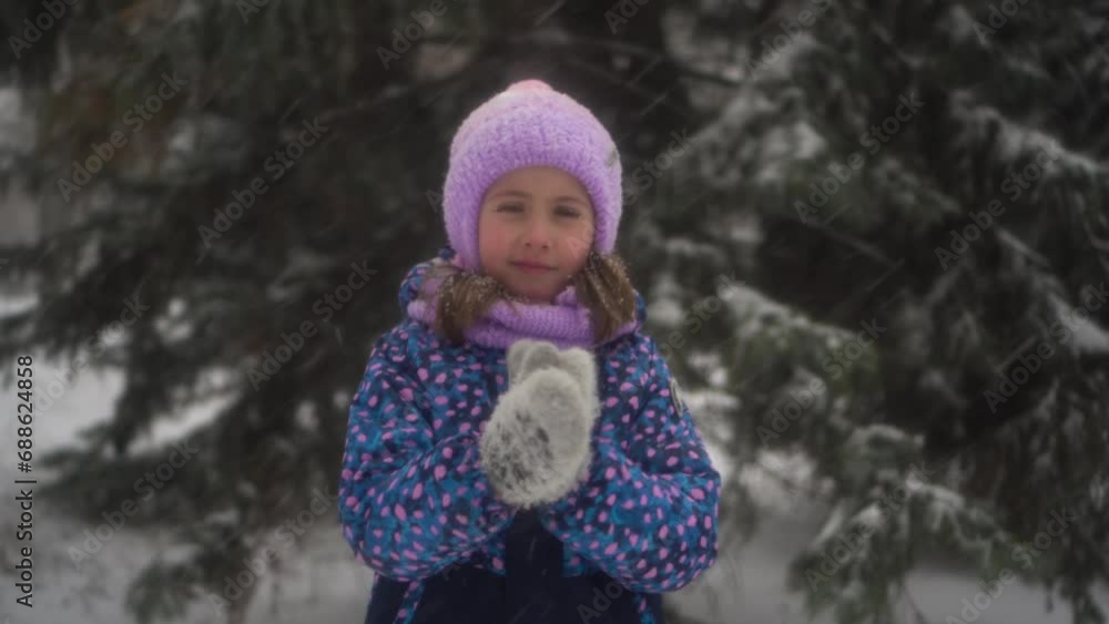 A girl stands against the backdrop of fir trees under snowfall and rubs her hands against the cold. A girl in a jacket and hat stands under heavy snowfall. A girl stands against the backdrop of fir trees under snowfall and rubs her hands against the cold. A girl in a jacket and hat stands under heavy snowfall.