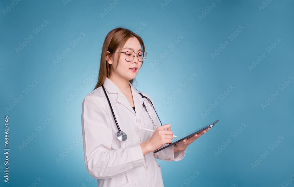 Young asian doctor wear medical uniform lab coat and stethoscope on blue background. Smiling female doctor writing diagnosis in digital tablet. Side view. Young asian doctor wear medical uniform lab coat and stethoscope on blue background. Smiling female doctor writing diagnosis in digital tablet. Side view.