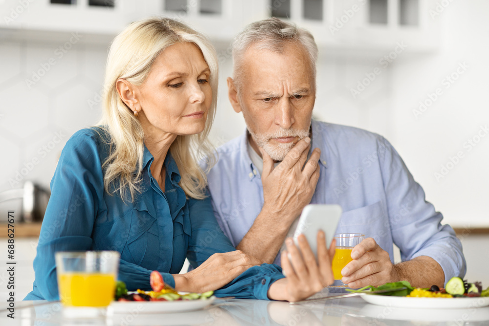 Worried Older Spouses Looking At Smartphone And Having Breakfast In Kitchen Worried Older Spouses Looking At Smartphone And Having Breakfast In Kitchen
