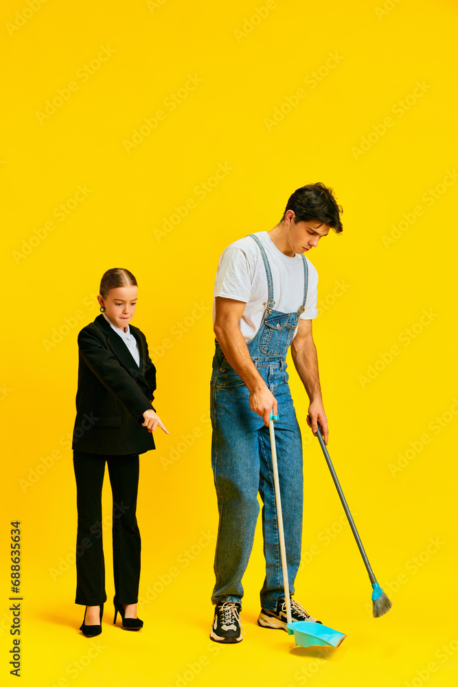 finicky manager, little girl, schoolgirl tells janitor where on floor to pick up dust on floor against vivid yellow background. finicky manager, little girl, schoolgirl tells janitor where on floor to pick up dust on floor against vivid yellow background.