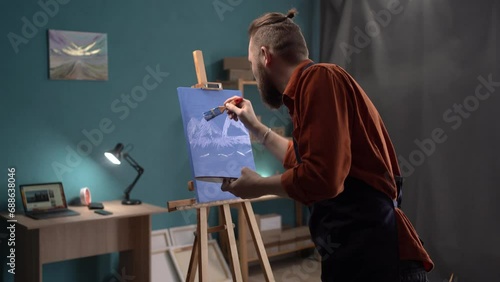 Artist painting mountains on a canvas with a paintbrush. Serious male painter making a blue painting for new art project. Creative man working in his art studio. photo