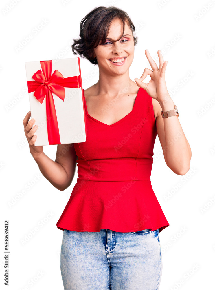 Beautiful young woman with short hair holding gift doing ok sign with fingers, smiling friendly gesturing excellent symbol Beautiful young woman with short hair holding gift doing ok sign with fingers, smiling friendly gesturing excellent symbol