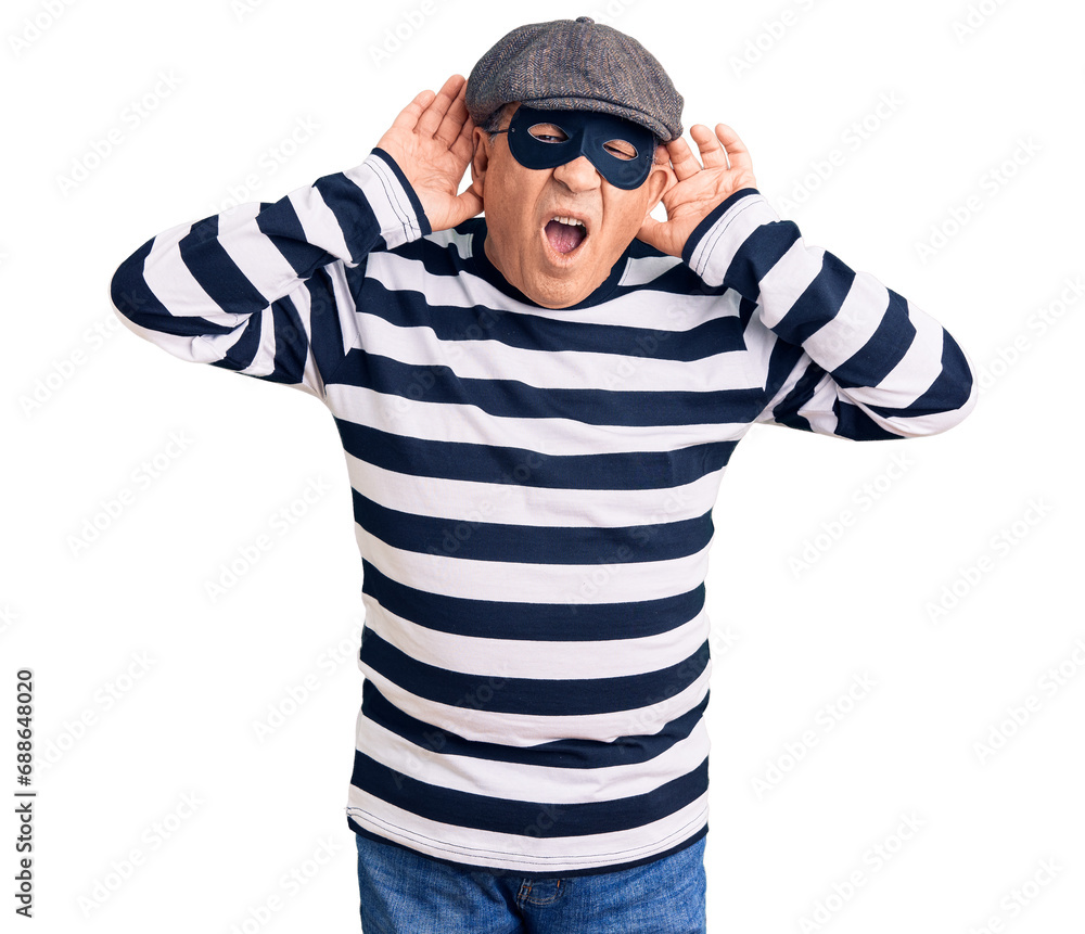 Senior handsome man wearing burglar mask and t-shirt trying to hear both hands on ear gesture, curious for gossip. hearing problem, deaf Senior handsome man wearing burglar mask and t-shirt trying to hear both hands on ear gesture, curious for gossip. hearing problem, deaf