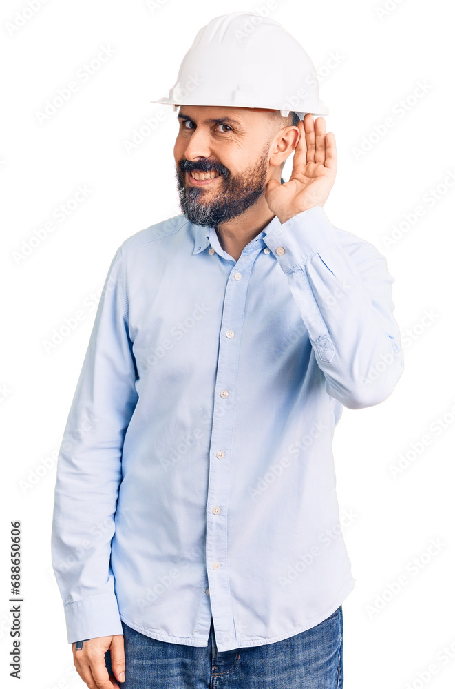 Young handsome man wearing architect hardhat smiling with hand over ear listening an hearing to rumor or gossip. deafness concept. Young handsome man wearing architect hardhat smiling with hand over ear listening an hearing to rumor or gossip. deafness concept.