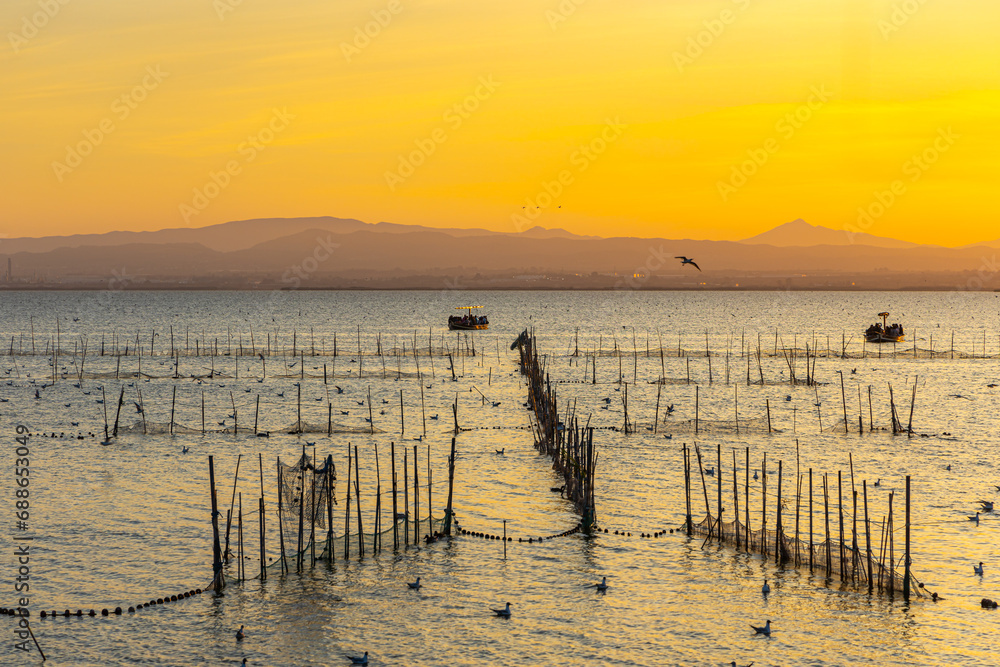 Obraz premium A boat with tourists and fishermen on a lake with fish nets and sunny path against the backdrop of an orange sunset