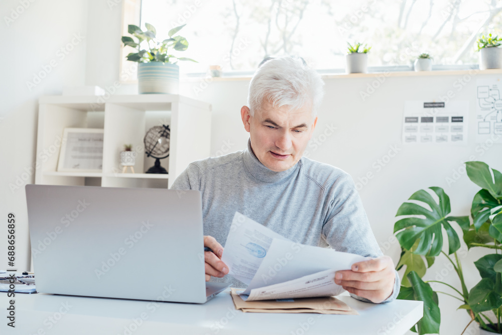 Middle aged businessman reading paper documents and using laptop while working at home office. Confident, experienced senior male professional. Small business entrepreneur manage business. paperwork Middle aged businessman reading paper documents and using laptop while working at home office. Confident, experienced senior male professional. Small business entrepreneur manage business. paperwork