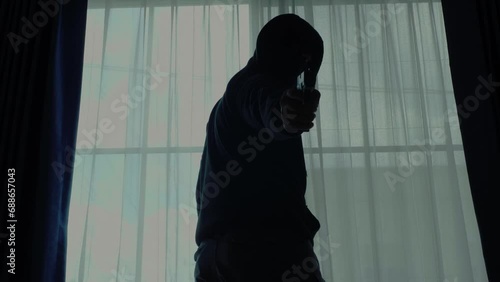 Mysterious man wearing black hoodie holding a pistol wearing a cloth mask covering his face completely in front of the door, Silhouette and dark concept image,threat of firearms,attack or defense.