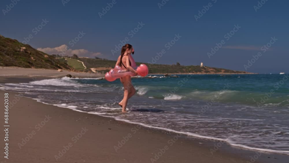 Woman walking on empty beach holding giant inflatable flamingo enjoying summer vacation on the beach wearing full one piece swimsuit. Woman having fun on summer travel vacation Woman walking on empty beach holding giant inflatable flamingo enjoying summer vacation on the beach wearing full one piece swimsuit. Woman having fun on summer travel vacation